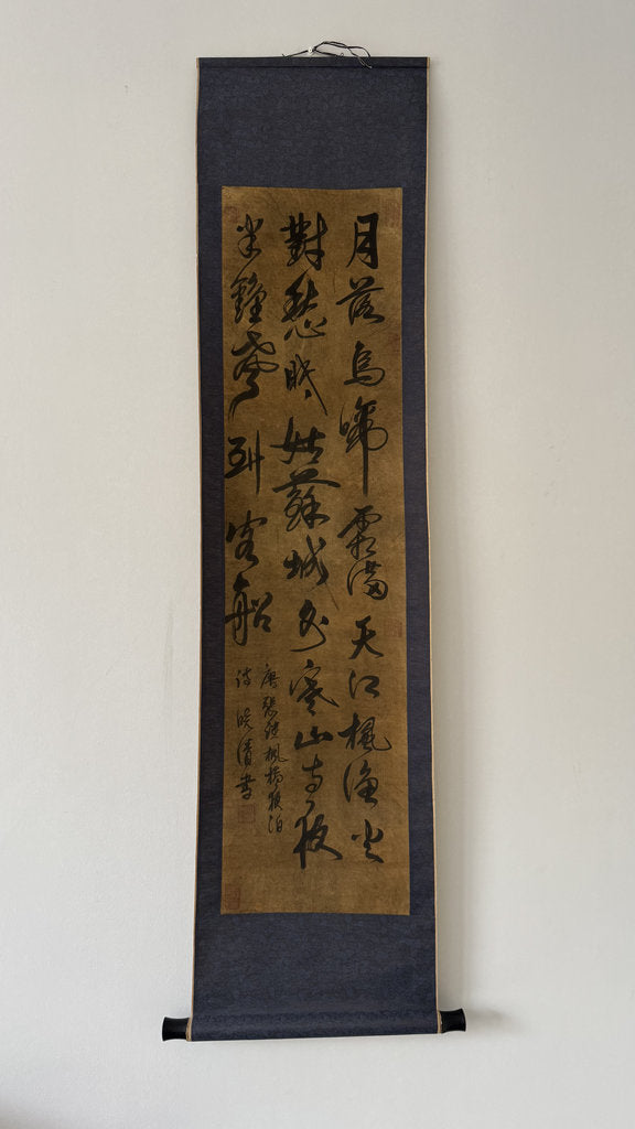 A traditional Japanse hanging calligraphy scroll with black ink script on a gold-toned paper center, mounted on a dark navy fabric border, shown hanging against a plain white wall.