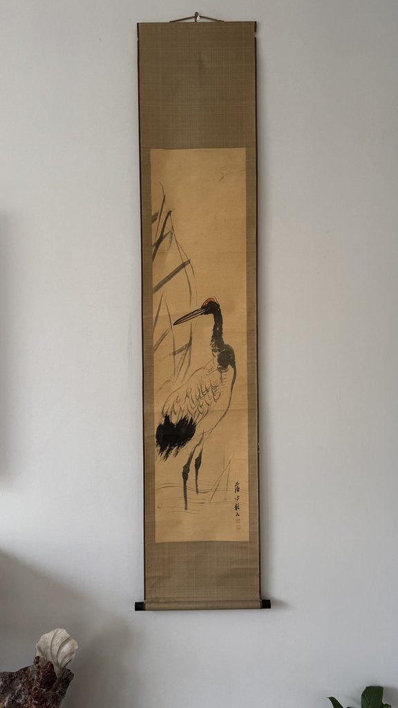 A traditional Japanese hanging scroll artwork featuring a black and white crane on a beige background, mounted on a light brown fabric wall hanging and displayed against a white wall.