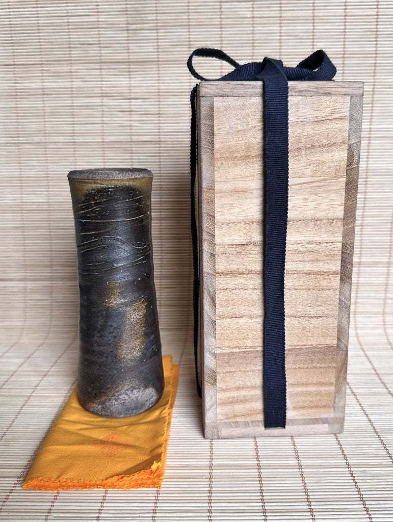 A dark textured ceramic vase with a flared rim sits on a small orange cloth next to a tall rectangular wooden presentation box tied with a dark ribbon, set against a bamboo mat background.