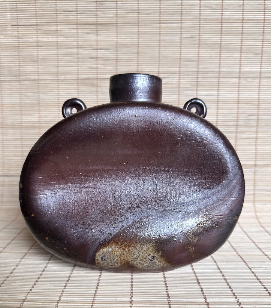 A dark brown ceramic flask with a rounded teardrop shape, a short neck, and two loop handles on the sides, resting on a light beige bamboo mat.
