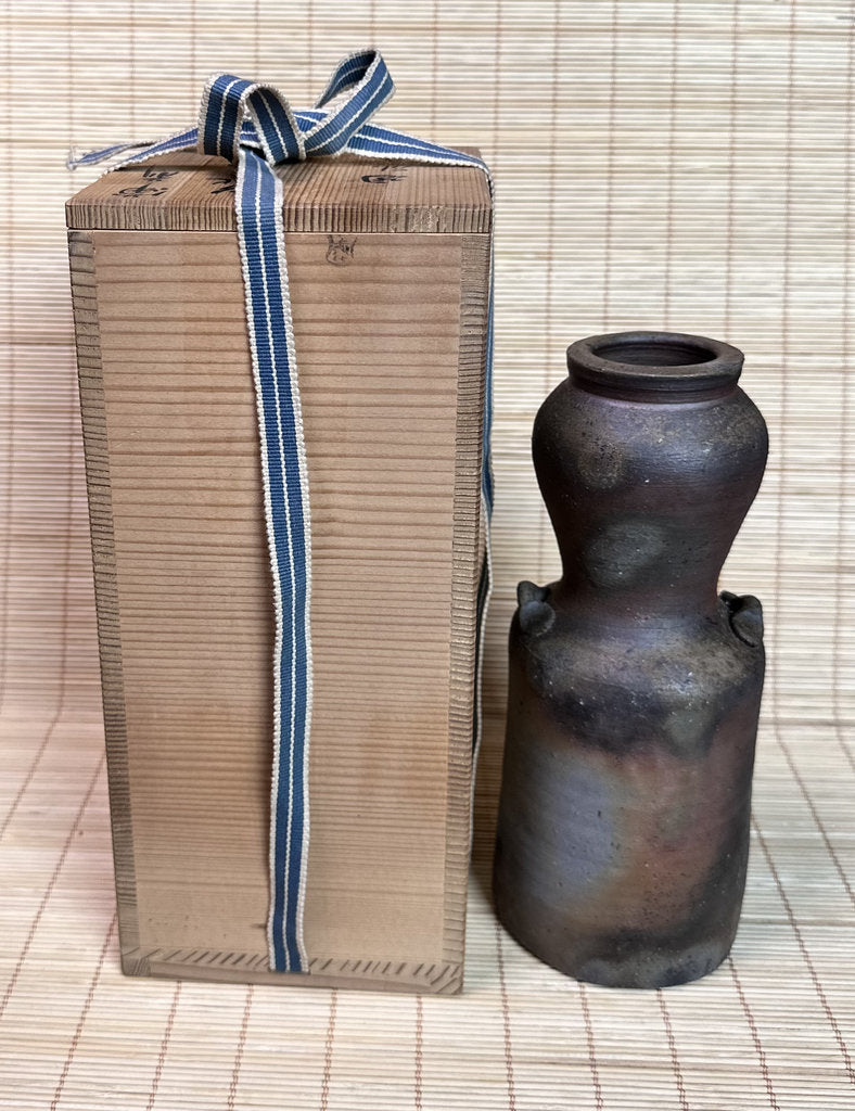 A dark brown ceramic vase with a matte, rustic finish stands next to its tall, brown cardboard gift box tied with a blue and white striped ribbon, set against a light beige woven mat background.