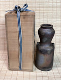 A dark brown ceramic vase with a matte, rustic finish stands next to its tall, brown cardboard gift box tied with a blue and white striped ribbon, set against a light beige woven mat background.
