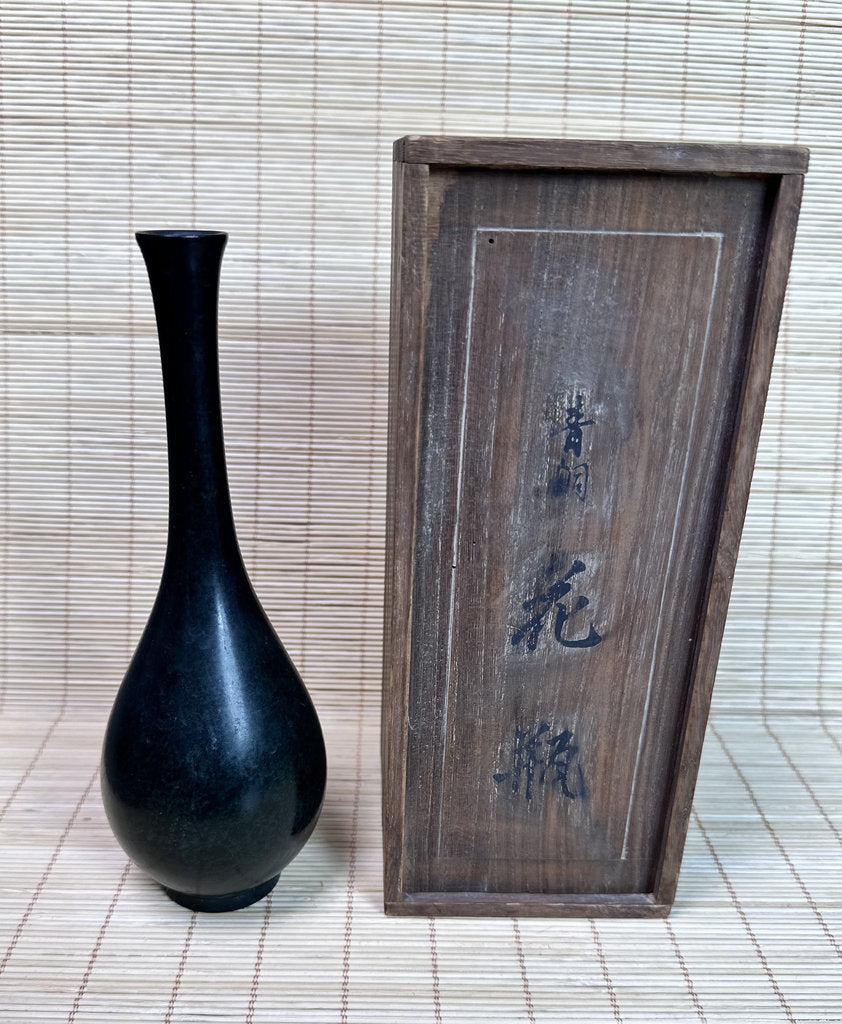 A glossy black ceramic vase with a long, slender neck and rounded body, displayed next to a rectangular dark wooden box with Japanese characters, set against a light beige woven mat background.