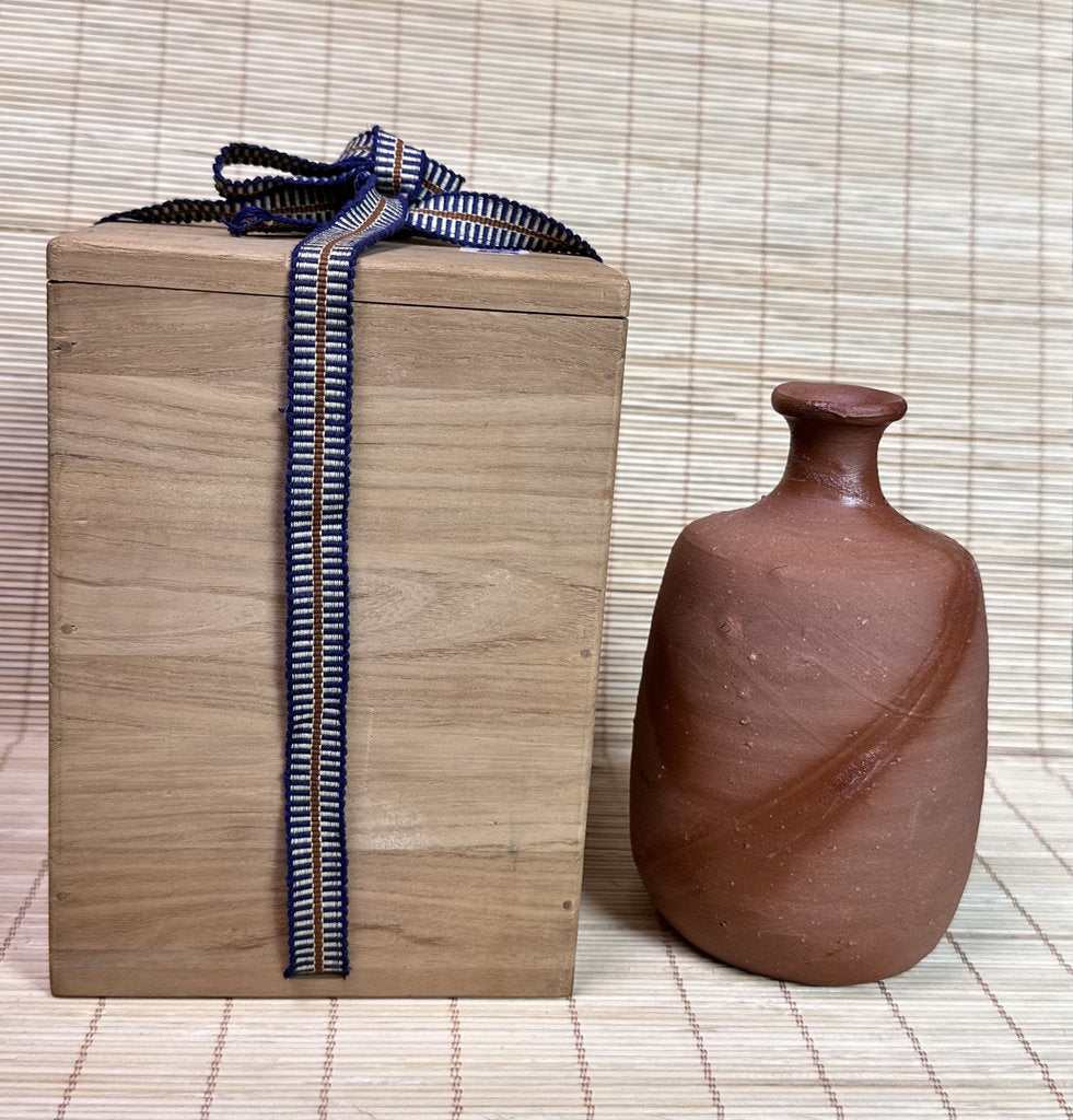 A small brown clay bottle with a narrow neck stands beside a light wood gift box tied with a navy and cream striped ribbon, set against a woven straw background.