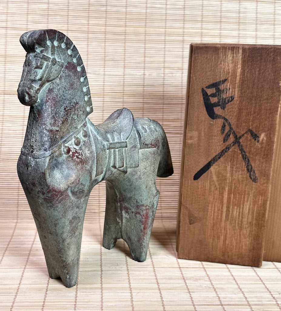 A small bronze horse figurine with a weathered patina stands next to a wooden box featuring a black engraved character, set against a light beige bamboo mat background.
