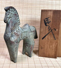 A small bronze horse figurine with a weathered patina stands next to a wooden box featuring a black engraved character, set against a light beige bamboo mat background.