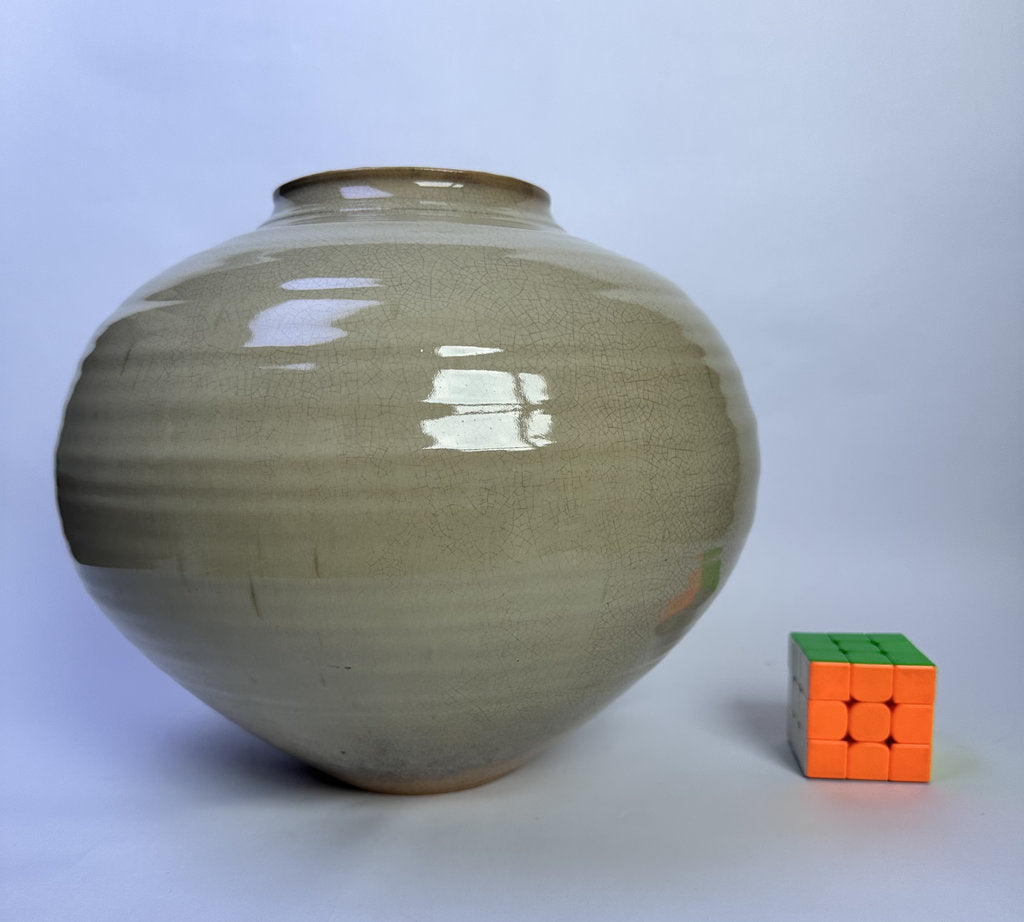A large round ceramic vase with a glossy, speckled beige-gray glaze, shown next to a small Rubik's cube for scale on a light gray background.