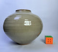 A large round ceramic vase with a glossy, speckled beige-gray glaze, shown next to a small Rubik's cube for scale on a light gray background.