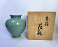 A glossy pale green ceramic vase with a flared rim and short pedestal base, shown next to a light wood box with black Japanese calligraphy on a plain white background.