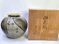 A round handmade ceramic vase with a textured gray and gold glaze, decorated with black floral patterns, shown next to its light brown wooden presentation box with black calligraphy.