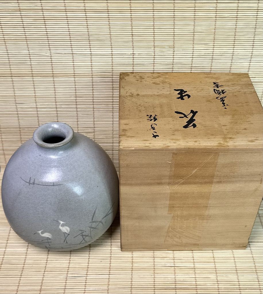 A small round gray ceramic vase with painted cranes and branches, placed beside a light natural wood presentation box with black calligraphy, set on a woven straw mat.