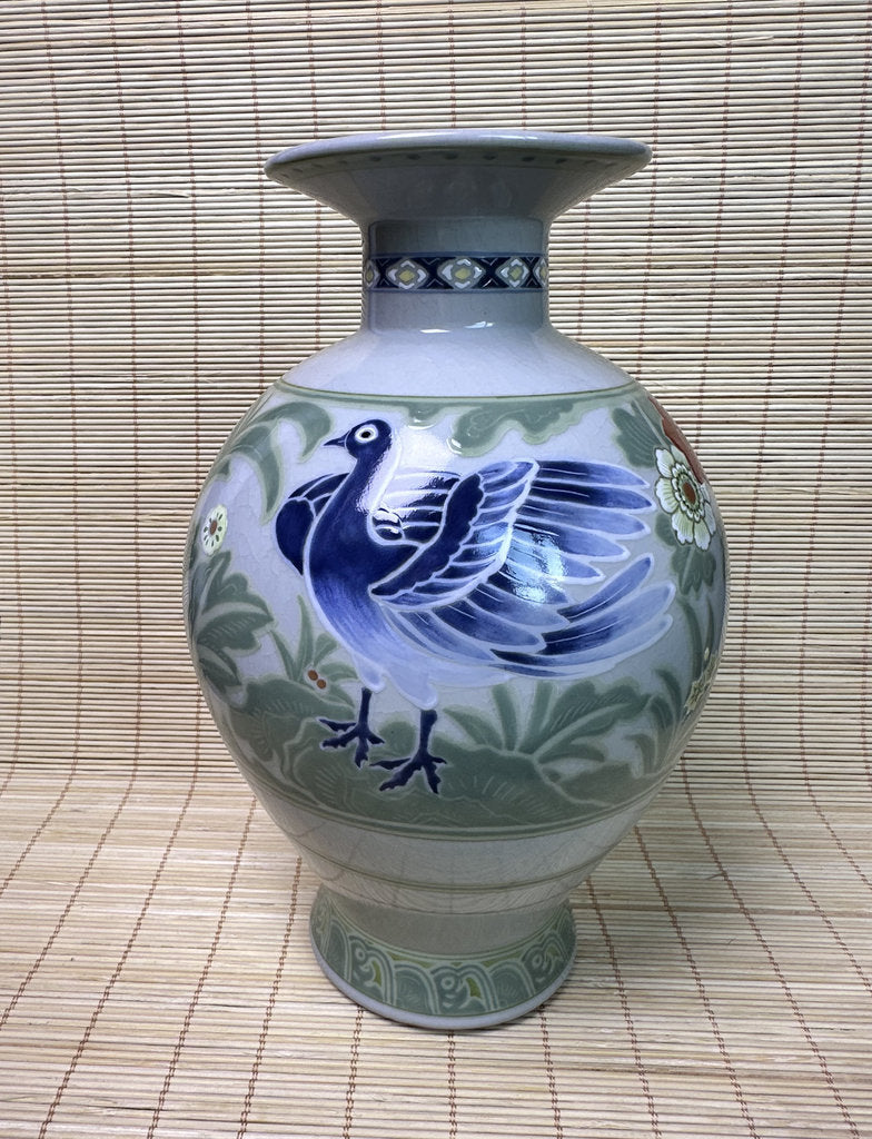 A ceramic vase with a flared rim and pedestal base, decorated with a blue bird and green floral motifs on a pale background, set against a woven straw mat backdrop.
