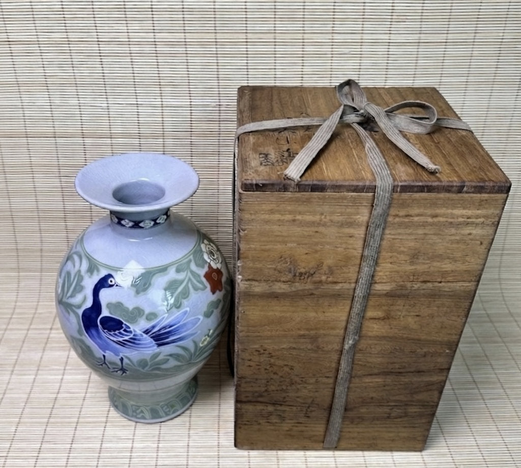 A decorative ceramic vase with a blue bird and green floral pattern next to a rustic wooden gift box tied with a gray ribbon, set against a bamboo mat background.