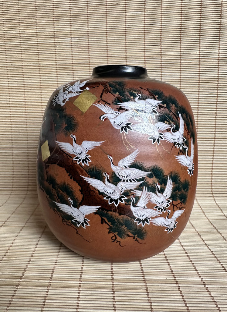 A brown ceramic jar decorated with hand-painted white cranes, black pine trees, and small gold accents, set against a woven straw mat background.