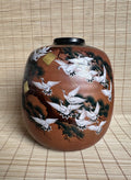 A brown ceramic jar decorated with hand-painted white cranes, black pine trees, and small gold accents, set against a woven straw mat background.