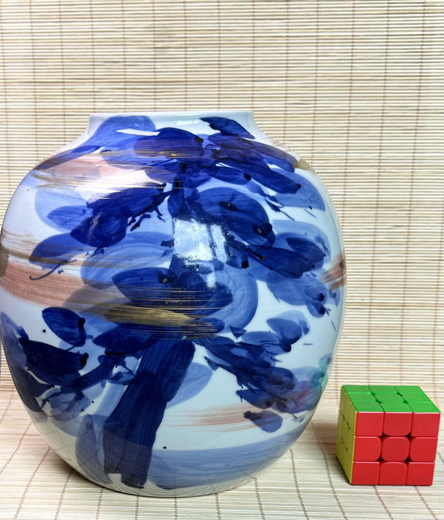 A round ceramic vase with a glossy white glaze decorated with expressive blue and brown brushstrokes, shown next to a small 3x3 Rubik's cube for scale on a light bamboo mat background.