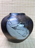 A ceramic bud vase with a glossy, mottled glaze transitioning from deep black and brown to light blue, decorated with delicate hand-painted gray branches, set against a light beige woven mat background.