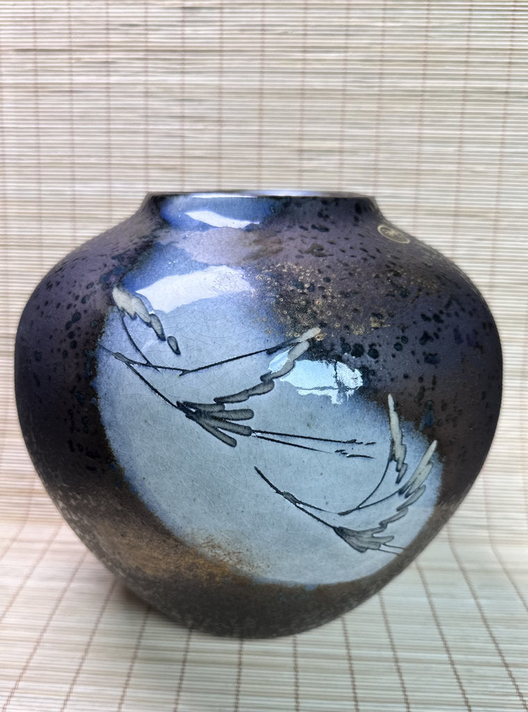 A ceramic bud vase with a glossy, mottled glaze transitioning from deep black and brown to light blue, decorated with delicate hand-painted gray branches, set against a light beige woven mat background.