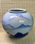A ceramic jar with a rounded body and wide rim, decorated with a hand-painted blue and beige mountain landscape design on a glossy white glaze, set against a bamboo mat background.