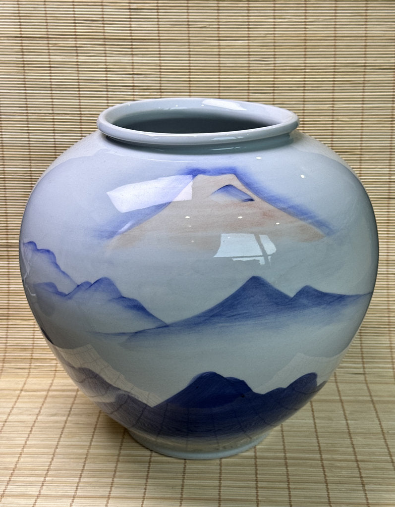 A ceramic jar with a rounded body and wide rim, decorated with a hand-painted blue and beige mountain landscape design on a glossy white glaze, set against a bamboo mat background.