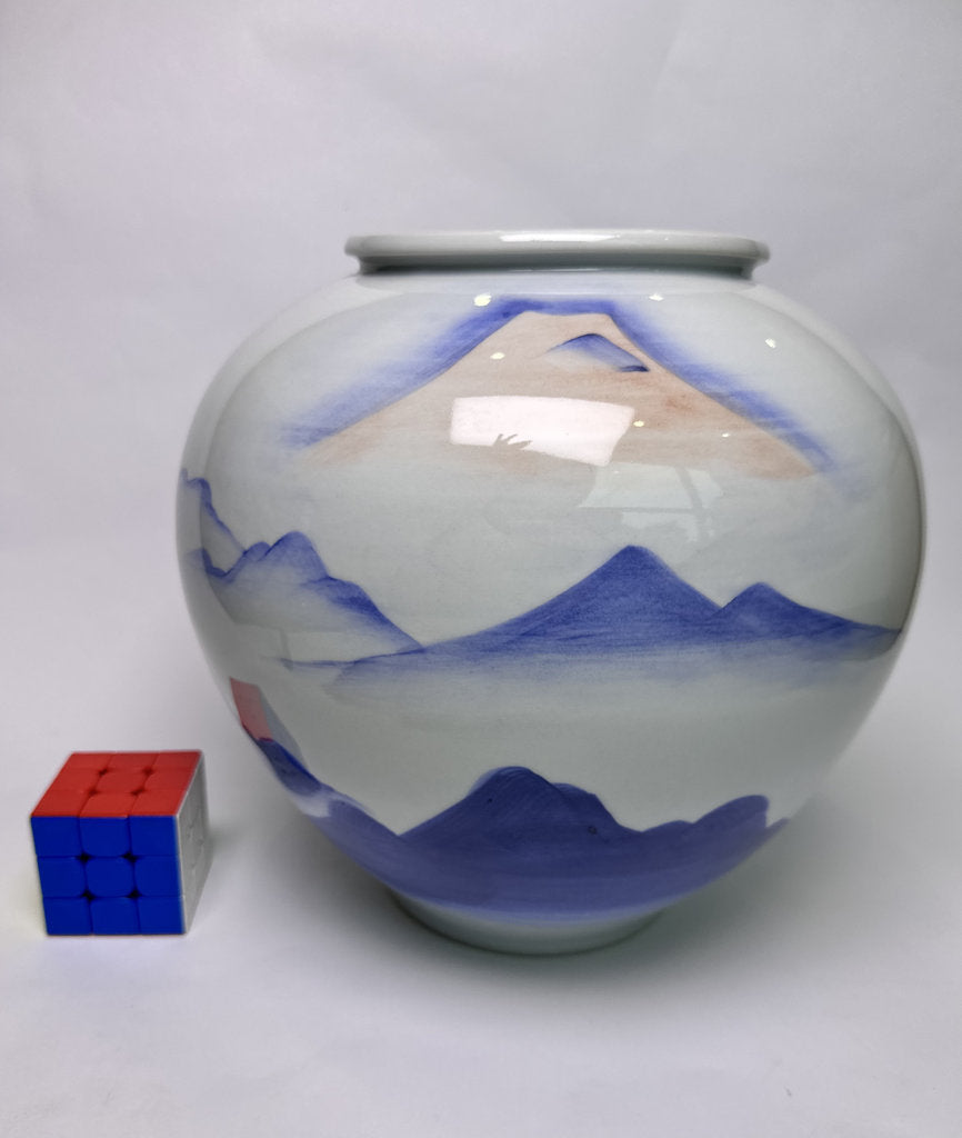 A white ceramic jar decorated with a blue and beige hand-painted mountain landscape, shown next to a small red, blue, and white puzzle cube for scale on a light gray background.